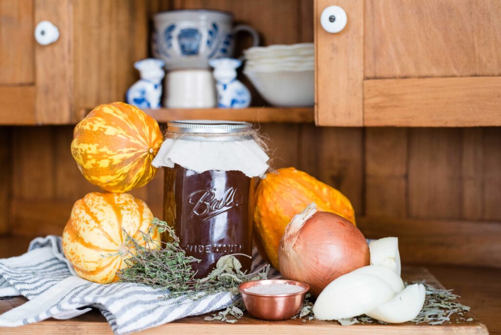 Jar of homemade bone broth with whole squash, onions, herbs and bowl of salt