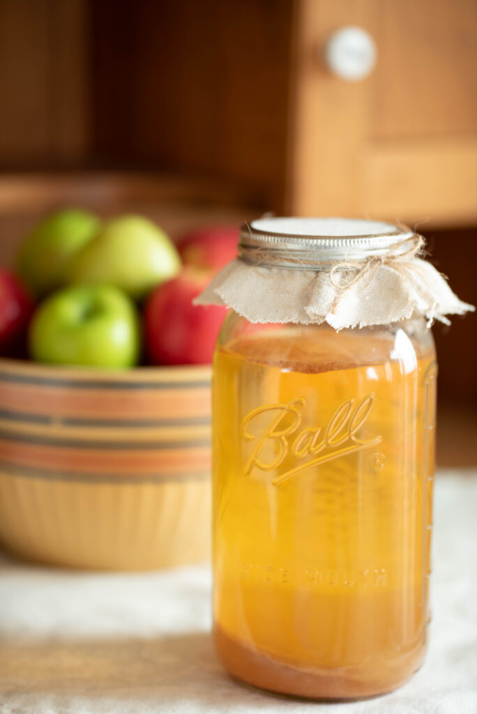 Jar of homemade apple cider vinegar with a bowl of fresh apples.