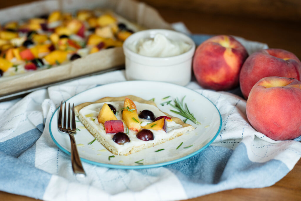 Piece of fruit pizza with bowl of vanilla bean ganache and whole peaches.