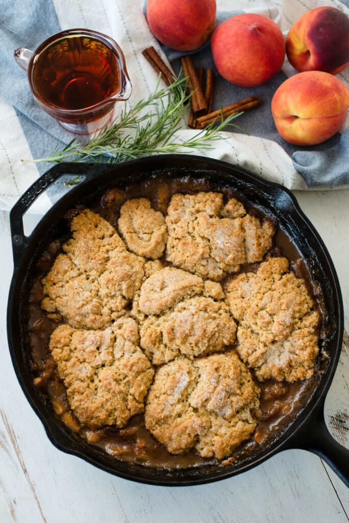 Fully baked old fashioned peach cobbler with saucer of maple syrup, fresh rosemary, cinnamon sticks and whole peaches.