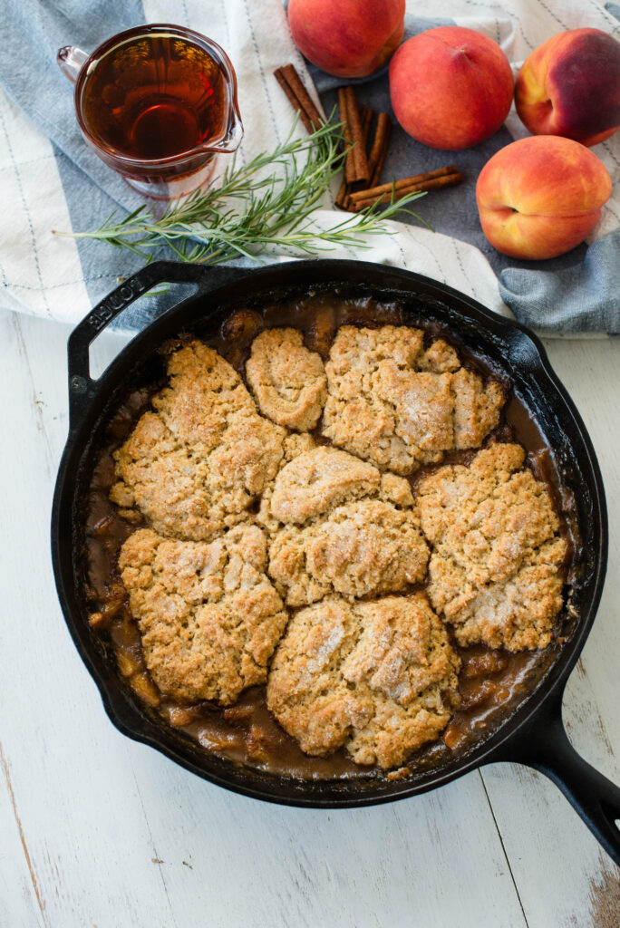 Old-fashioned peach cobbler in a skillet with fresh peaches, rosemary, cinnamon sticks and saucer of maple syrup.