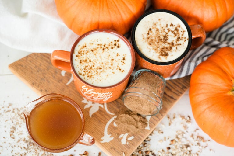 Two mugs of herbal pumpkin spice latte pictured with fresh whole pumpkins, herbs and pumpkin spice mix.